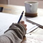 Background Check - person writing on brown wooden table near white ceramic mug