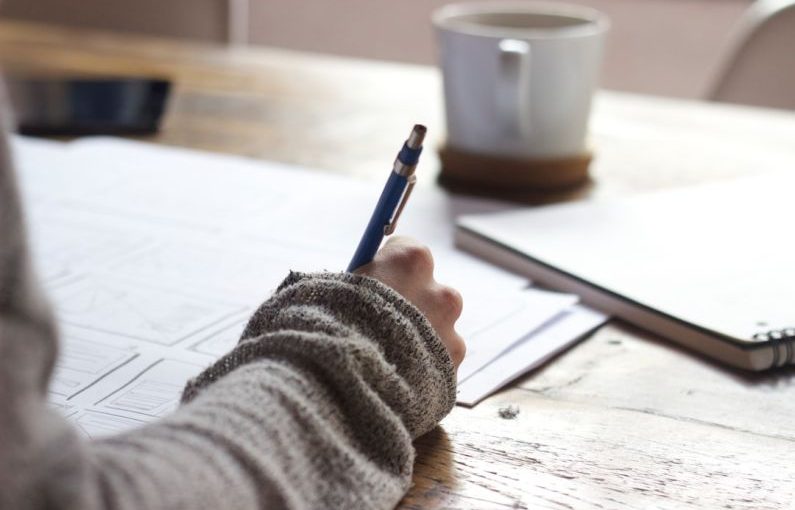 Background Check - person writing on brown wooden table near white ceramic mug